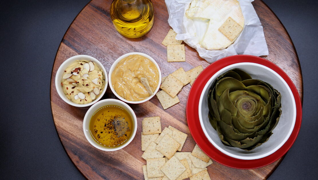A wooden board with cheese, crackers and artichokes.